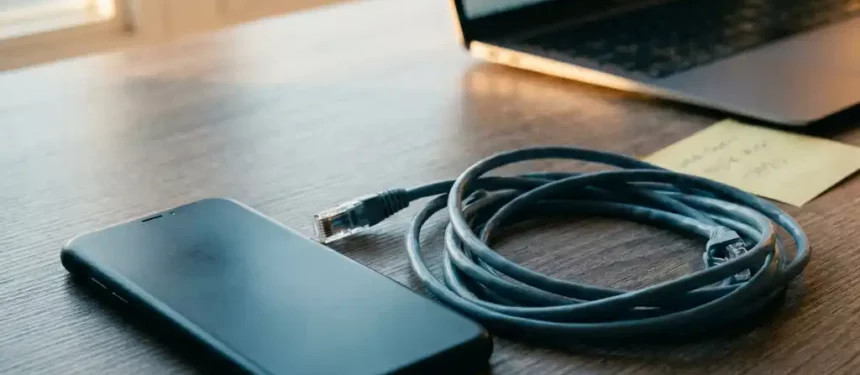 Smartphone met scherm naar beneden op bureau naast loshangende ethernet-kabel, sticky note en laptop op de achtergrond.